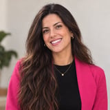 Woman with long dark wavy hair smiling, wearing a bright pink blazer over a black top and a gold pendant necklace
