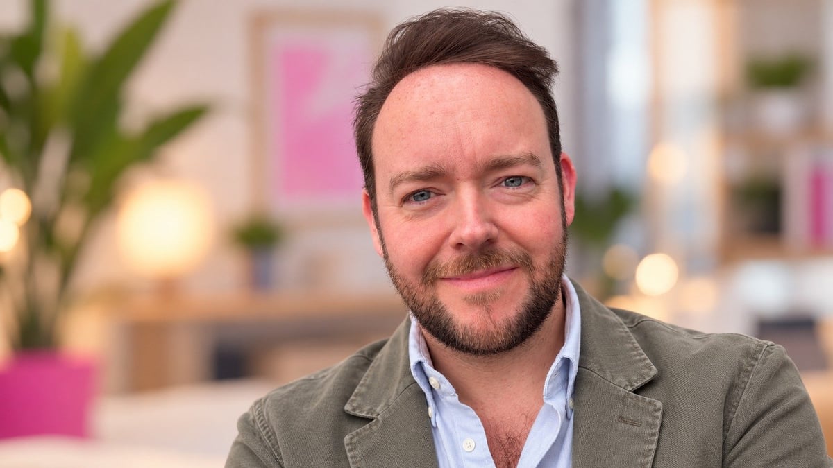 Man with dark hair and beard wearing gray blazer and light blue shirt, smiling in modern office setting